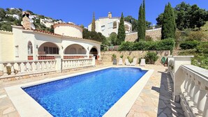 A swimming pool with a stone deck, a white building with arches, and a stone wall with greenery.