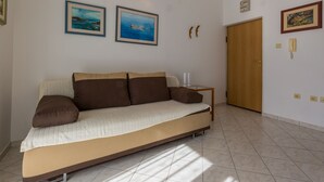 Une chambre avec un lit simple, une porte en bois, deux tableaux encadrés au mur, une table de chevet avec une lampe et un sol carrelé.