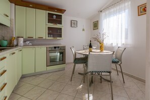 Une cuisine avec des meubles de couleur vert clair, une table à manger avec des chaises, une horloge au mur et une fenêtre avec des rideaux.