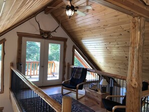 A cozy cabin interior with a wooden ceiling, a ceiling fan, a fireplace, a large window, and a balcony. - Bear Hideaway (Packwood)
