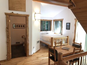 Una habitación con estructura de madera, una cama, una mesa y una silla. Hay una ventana con cortinas y un cuadro en la pared.