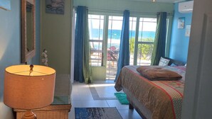 A bedroom with a bed, a lamp, a wicker chair, a view of the sea through the window, and a ceiling fan.