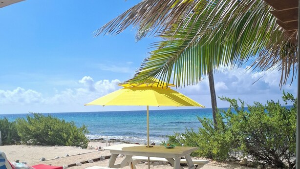 A beach view with a yellow umbrella, a wooden table, and a white lounge chair with a red cushion.