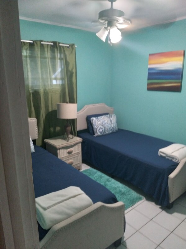 A bedroom with two beds, a nightstand, a lamp, a ceiling fan, and a painting on the wall.