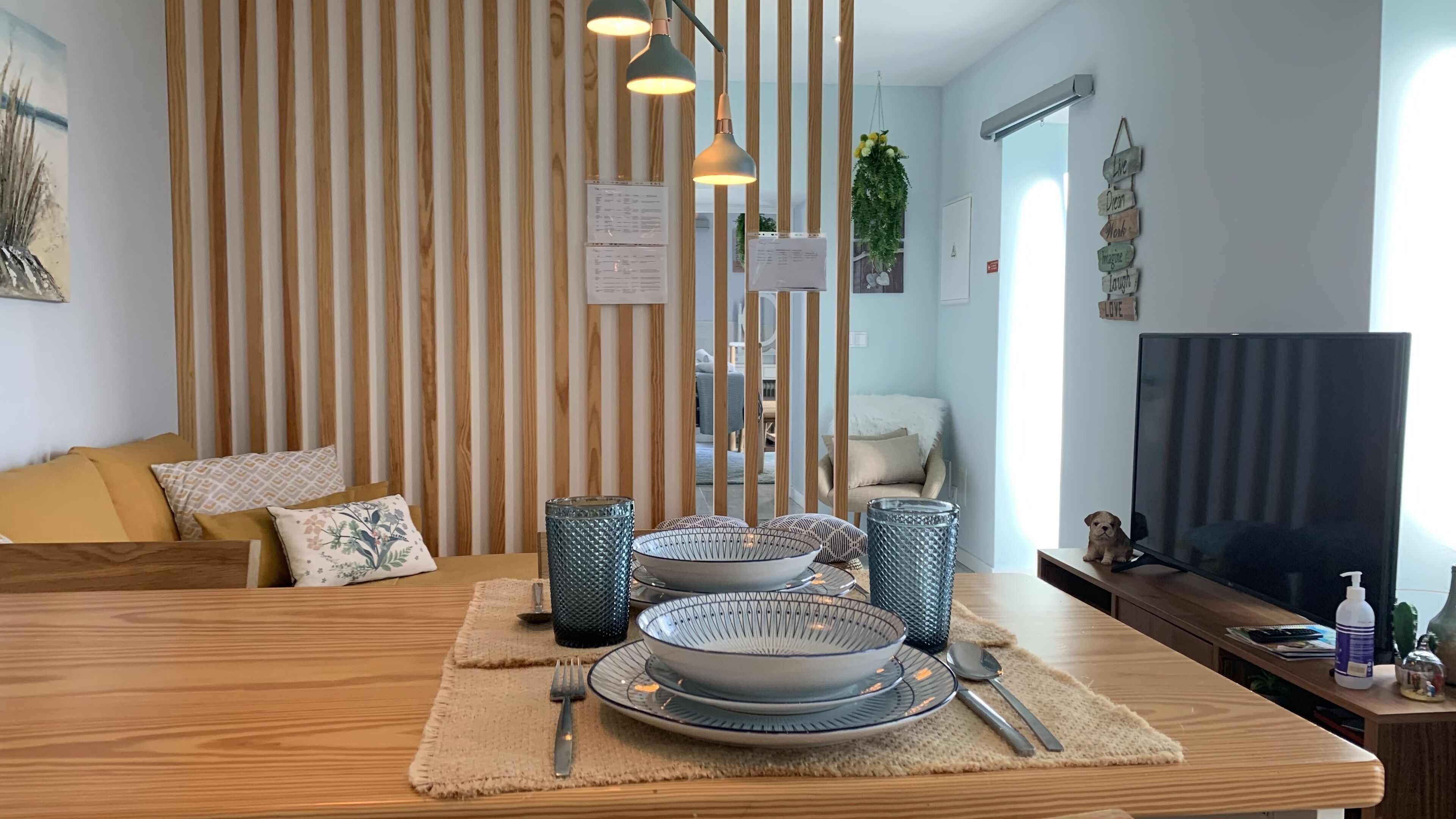 Un salon moderne avec une table à manger en bois, dressée pour deux personnes, un canapé avec des coussins, un téléviseur et un mur décoratif orné de lattes de bois verticales.
