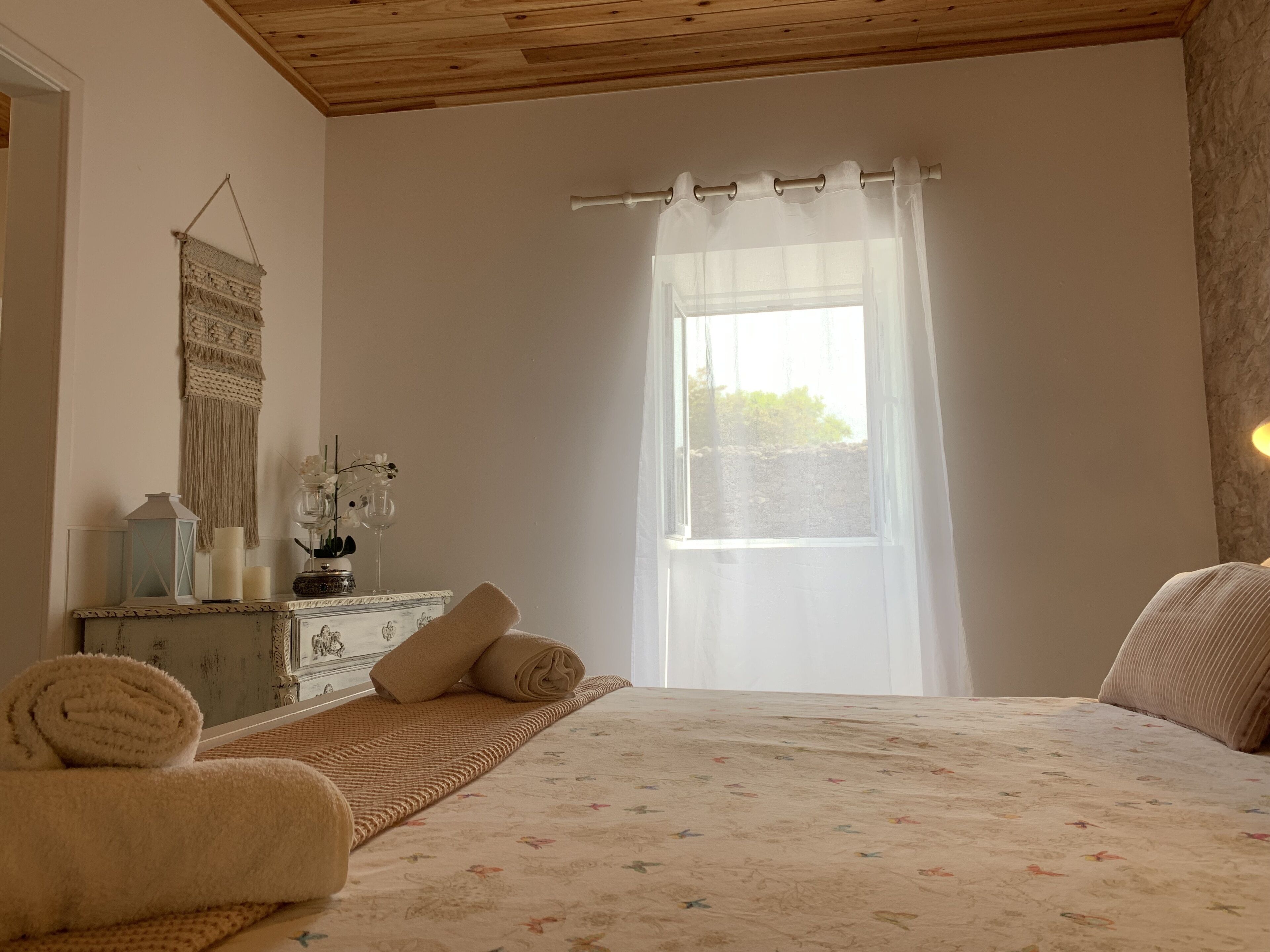 Un dormitorio con cama, una mesita de noche, un espejo, una ventana con cortinas translúcidas y un adorno de macramé en la pared.