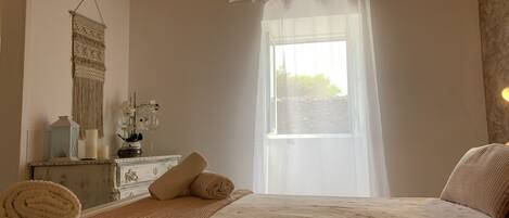 Un dormitorio con cama, una mesita de noche, un espejo, una ventana con cortinas translúcidas y un adorno de macramé en la pared.