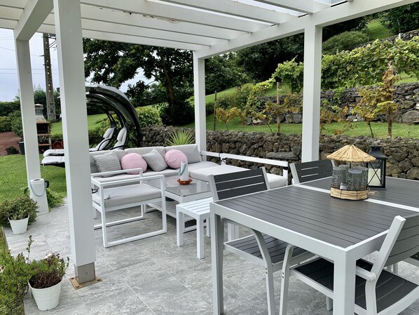 Une terrasse extérieure agrémentée d’une pergola blanche, d’une table ronde avec des chaises, d’un canapé avec des coussins et de plantes en pot.