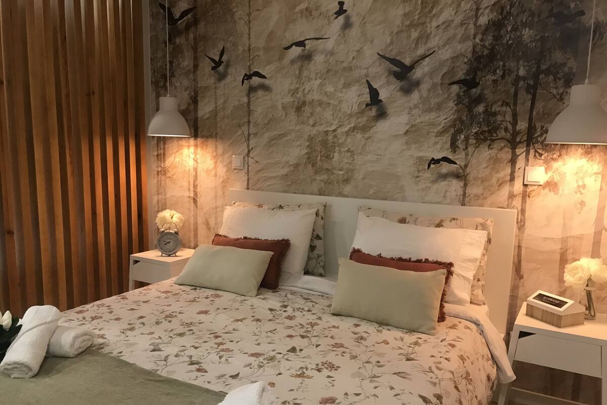 A bedroom with a large bed, two bedside tables, a wall clock, and a wall-mounted light fixture.