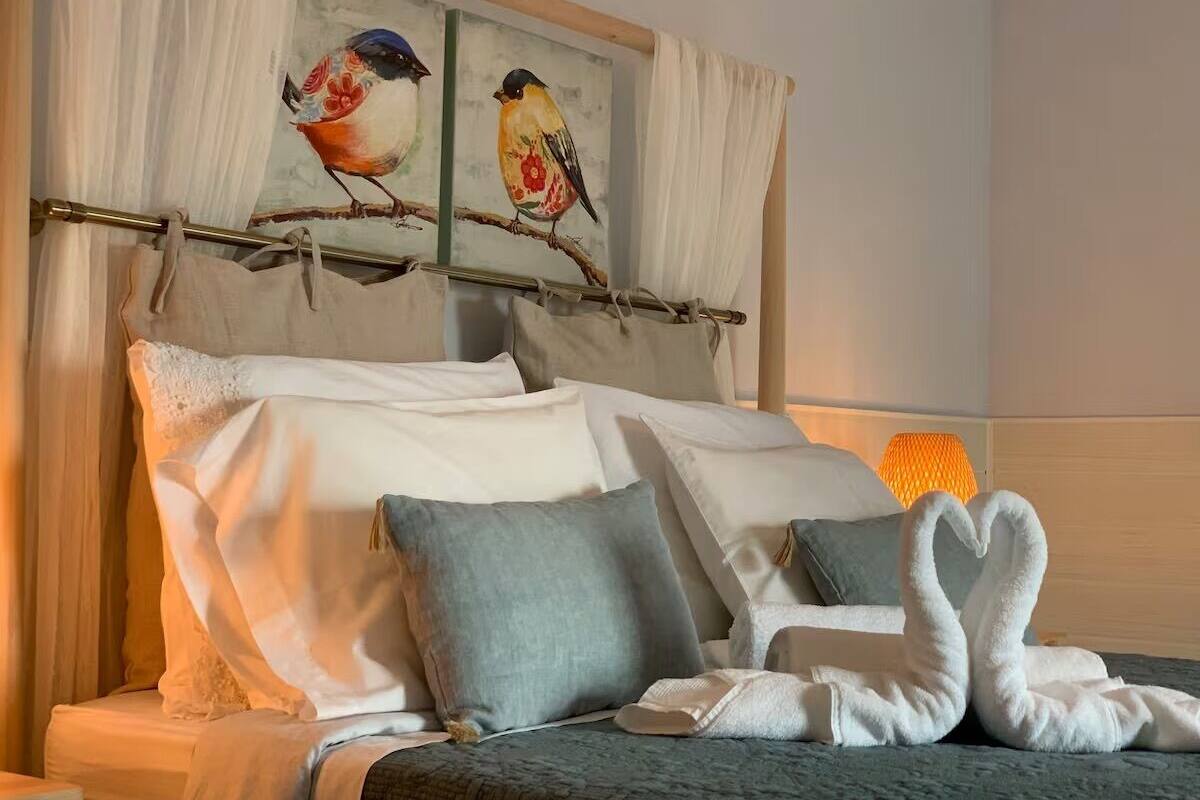 A neatly made bed with white and grey linens, a pair of white towels arranged in a heart shape, and a painting of birds on the headboard.