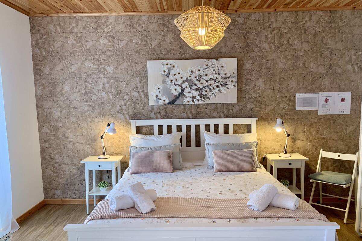 A bedroom with a stone wall, a wooden ceiling, a white bed with floral bedding, two bedside tables with lamps, and a chair.