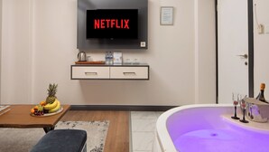 A modern living room with a flat-screen TV displaying 'NETFLIX', a wooden table with fruit, a sofa, and a jacuzzi tub with champagne glasses. - Bakırköy Zeugma Suit  (Istanbul)