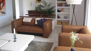 A modern living room with a brown leather sofa, a white coffee table, a bookshelf, and a floor lamp. - Ref. Nav44 -  (Rosas / Roses)