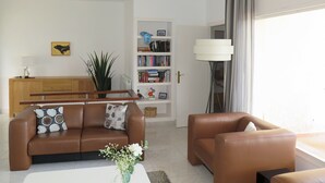 A modern living room with a brown leather sofa, a white coffee table with a vase of flowers, a bookshelf, and a lamp near a window. - Ref. Nav44 -  (Rosas / Roses)