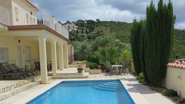 A swimming pool with a tiled deck, a covered seating area with chairs, and a view of a hillside with buildings. - Ref. Nav44 -  (Rosas / Roses)