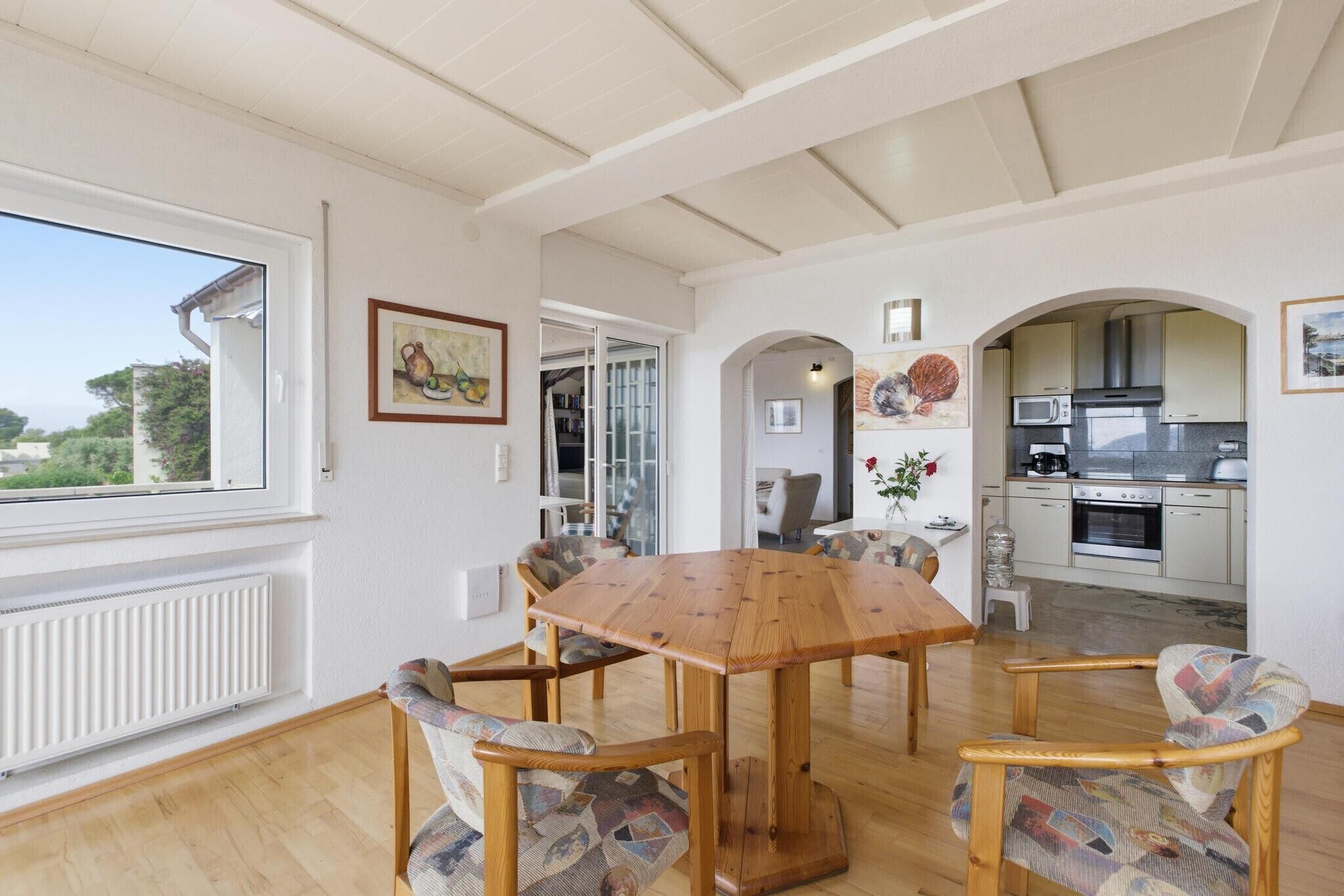 A dining area with a wooden table and chairs, a kitchen in the background, and a view through a window.