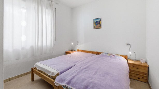 A bedroom with a double bed, two bedside tables, a nightstand, a wall-mounted lamp, and a painting.
