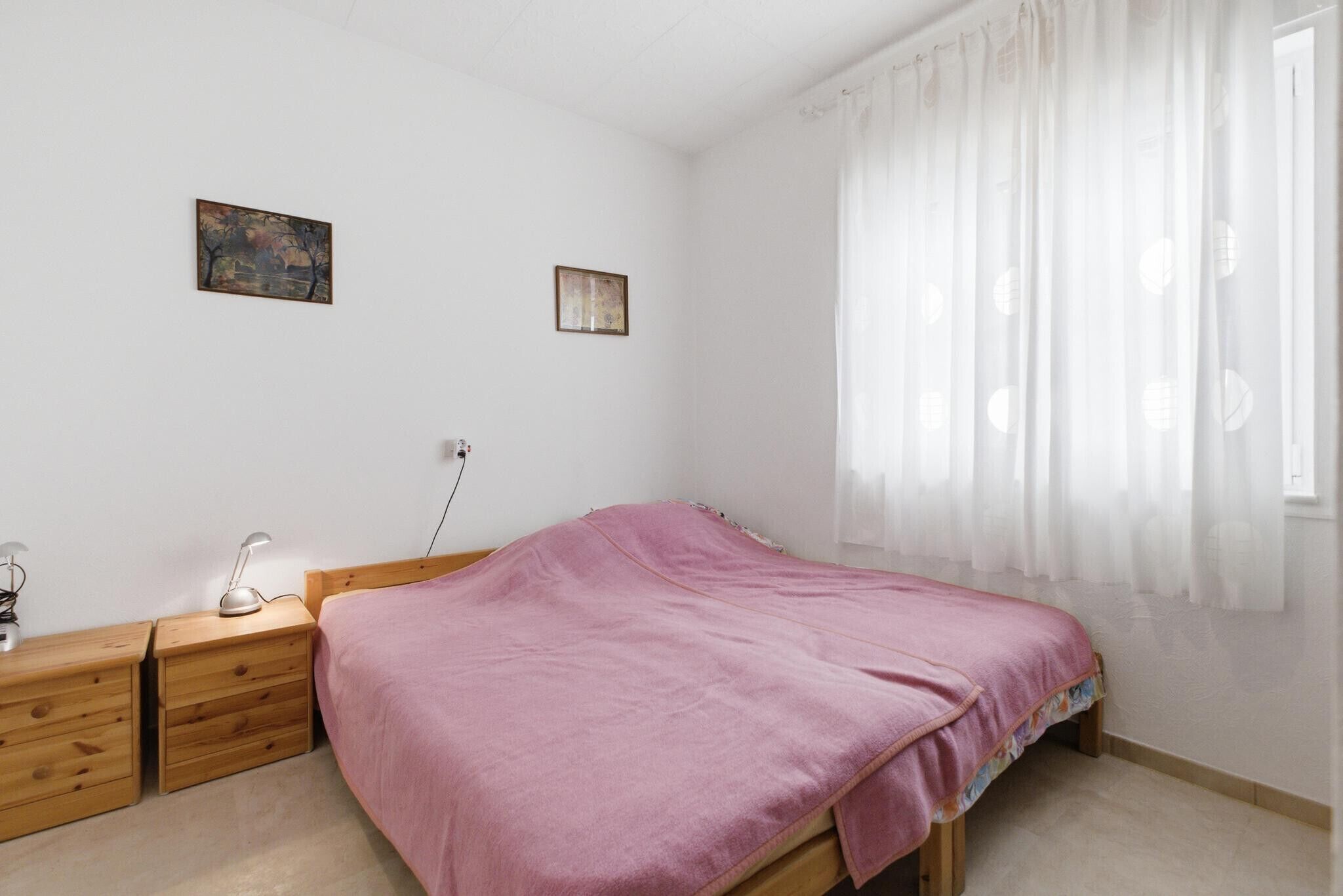 Un dormitorio con cama, mesitas de noche de madera, un escritorio, una lámpara y dos cuadros colgados en la pared.