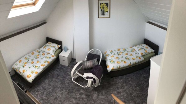 Una habitación compacta con dos camas individuales, una mesita de noche pequeña y blanca, una silla de mimbre y un cuadro enmarcado en la pared.
