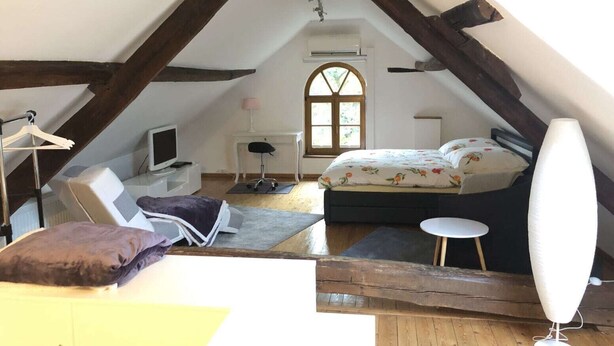Une chambre mansardée chaleureuse, avec un lit, un bureau, une chaise, des poutres en bois et une fenêtre.