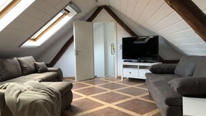 Une chambre confortable située sous les toits, avec un plafond en pente, une grande fenêtre, un téléviseur à écran plat et deux canapés gris.