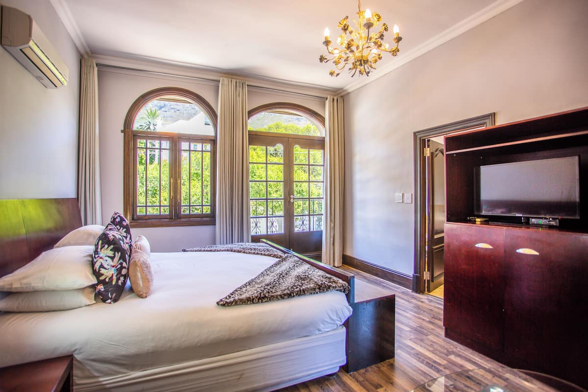 Une chambre à coucher avec un grand lit, deux fenêtres avec des rideaux, un téléviseur posé sur un meuble en bois et un lustre.