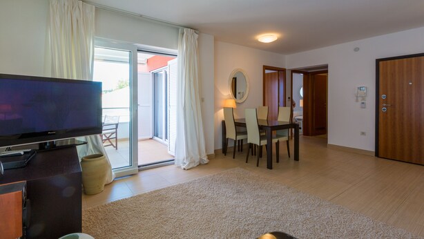 Uma sala de estar moderna com uma TV de tela plana, um espaço de refeições com mesa e cadeiras, uma janela grande com cortinas e uma porta de vidro deslizante que dá para uma área externa.