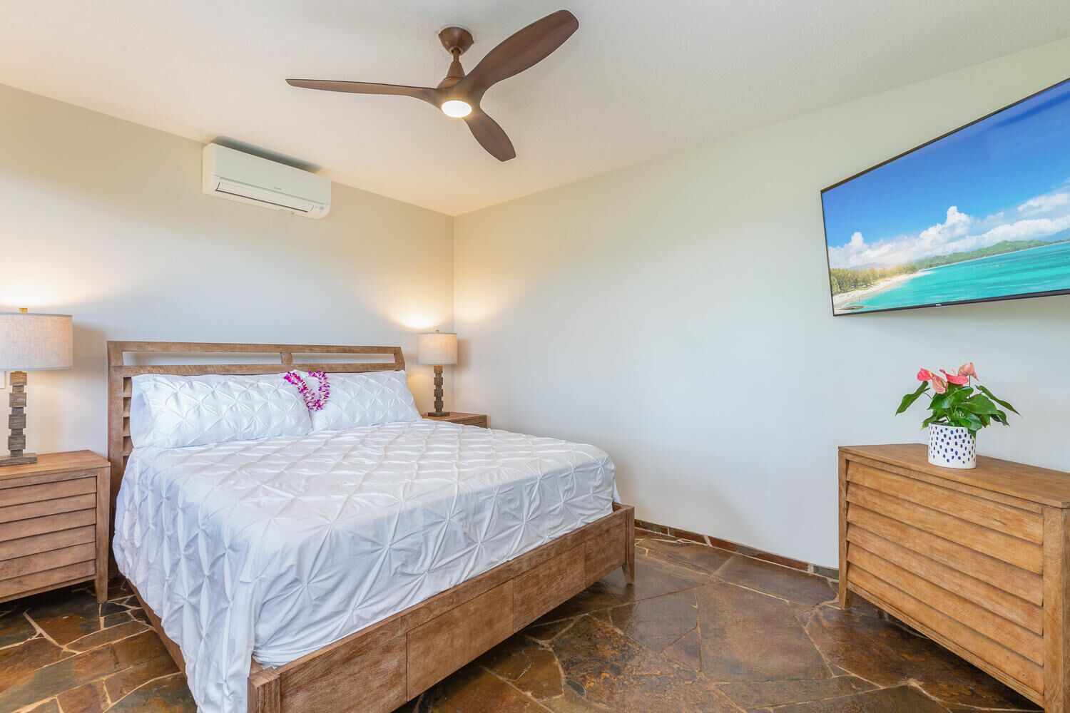 Un dormitorio con cama de madera, ventilador de techo, aire acondicionado instalado en la pared, mesita de noche con lámpara, cómoda de madera y un cuadro de una escena de playa.