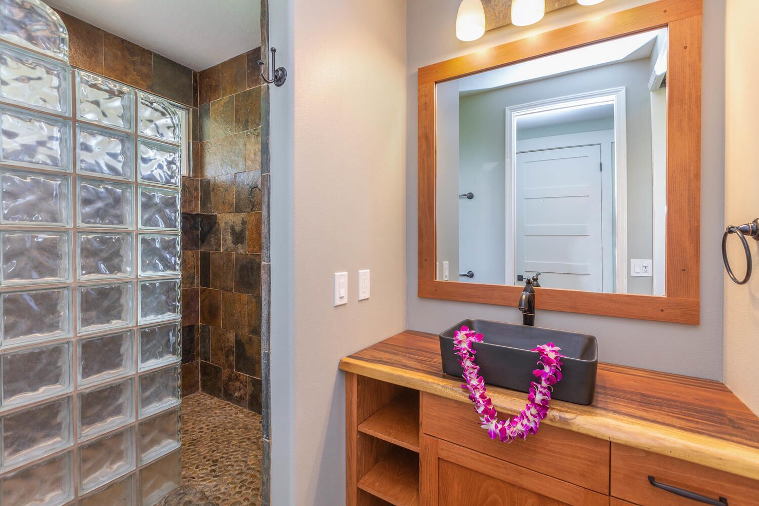 A bathroom with a glass block shower enclosure, a wooden vanity with a mirror, and a decorative lei.
