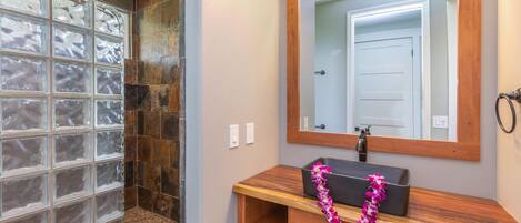 A bathroom with a glass block shower enclosure, a wooden vanity with a mirror, and a decorative lei.