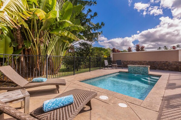 An outdoor swimming pool with lounge chairs, a stone wall, and tropical plants. - Pohaku Villa: Gorgeous Home w/AC, Pool, Relaxing Lanai Area & Garden Views (Princeville)