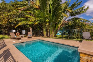 A swimming pool with lounge chairs and a lifeguard chair, surrounded by palm trees and a fence. - Pohaku Villa: Gorgeous Home w/AC, Pool, Relaxing Lanai Area & Garden Views (Princeville)