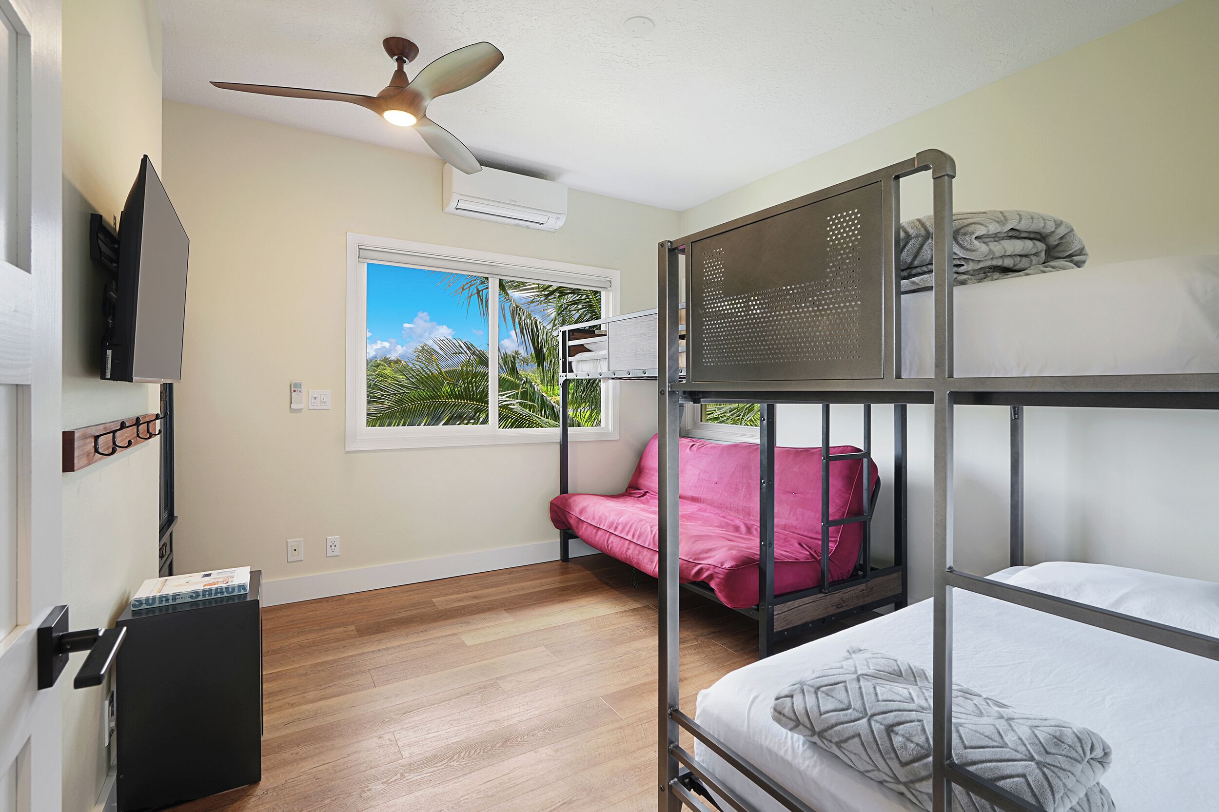 Une chambre avec un lit superposé, un ventilateur de plafond, une fenêtre donnant sur des palmiers et un petit bureau avec une chaise.