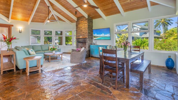 A spacious living area with a stone floor, wooden ceiling beams, a fireplace, and large windows offering a view of the outdoors.