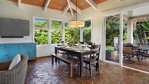 A spacious dining area with a stone floor, wooden ceiling beams, and large windows offering a view of lush greenery.
