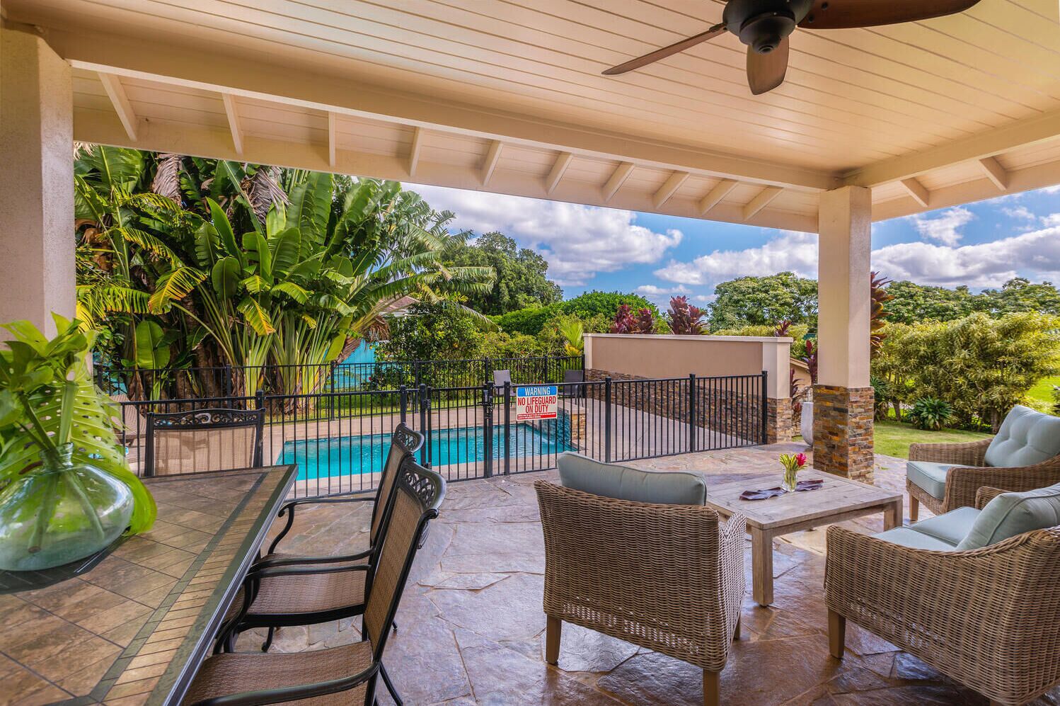 A covered outdoor area with a pool, wicker furniture, and lush greenery.