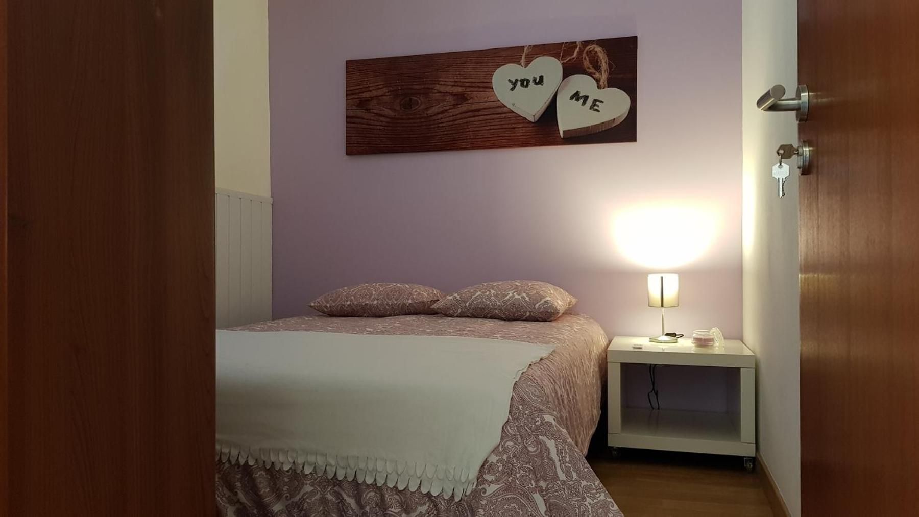 A bedroom with a bed, a nightstand, a wall-mounted heart-shaped decoration with 'you' and 'me' on it, and a lit lamp.