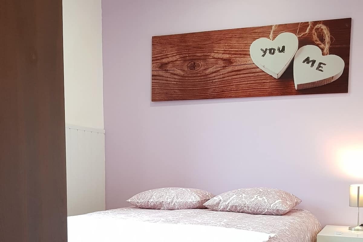 A bedroom with a bed, a nightstand, a lamp, a purple rug, and a wall decoration with hearts and the words "you" and "me".