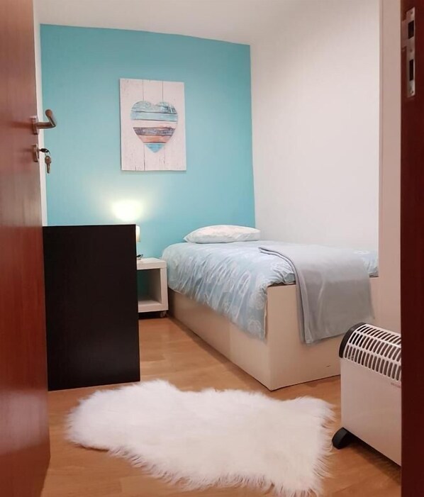A bedroom with a bed, a nightstand, a white furry rug, a heater, and a wall with a painting.