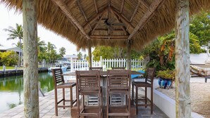 A covered outdoor seating area with a table and chairs, a thatched roof, and a view of a water body and white fence in the background. - Manatee Oasis 4bed/3bath single family home with private pool & dockage located on the gulf side of Marathon (Marathon)