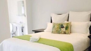 A neatly made bed with white bedding and a green throw blanket, a bedside table with a lamp, and a bathroom visible through an open door. - Red Dot Lodge (Randburg)