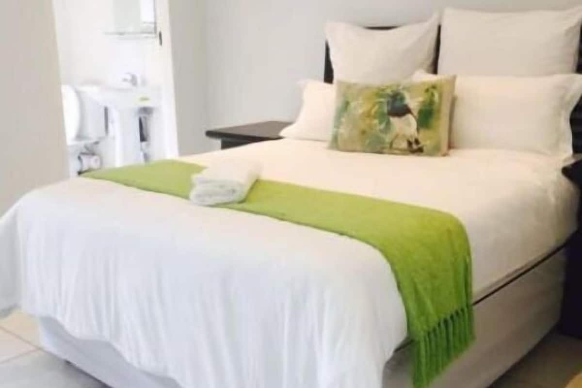 Una cama bien hecha con ropa de cama blanca y una manta verde, una mesita de noche con una lámpara, y un baño visible a través de una puerta abierta.