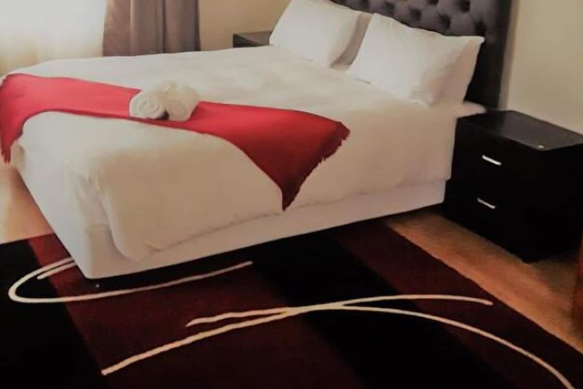 Una cama bien hecha con una colcha roja y almohadas blancas, un cabecero oscuro y capitonado, y una mesita de noche negra con cajones.