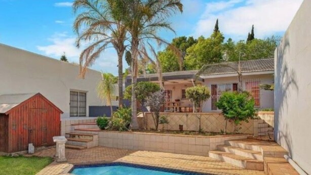 A swimming pool with a tiled deck, a red shed, and a covered patio area with a table and chairs. - Red Dot Lodge (Randburg)