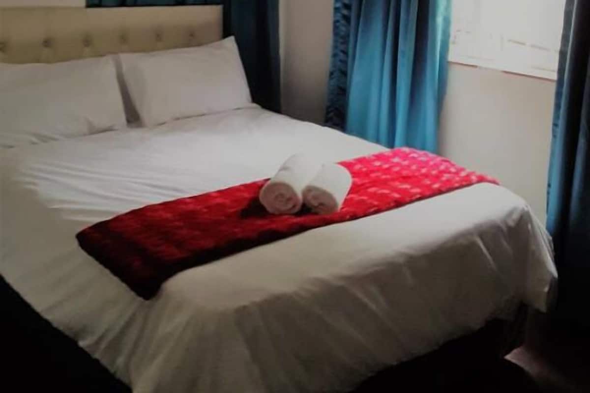 Una habitación de hotel con cama, ropa de cama blanca, una colcha roja y cortinas azul oscuro.