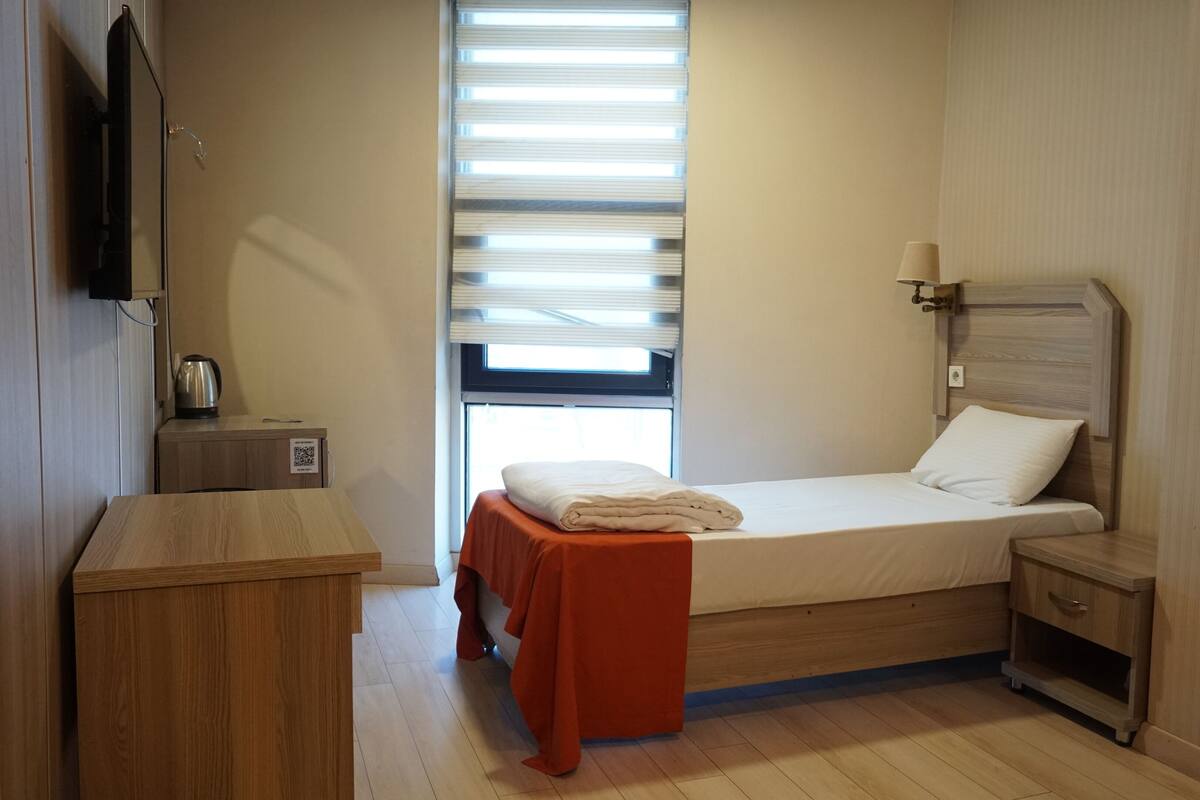 A small, neatly arranged room with a bed, a nightstand, a table, and a window with blinds.