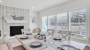 Un comedor con chimenea de ladrillo, paredes blancas y ventanas grandes con persianas.