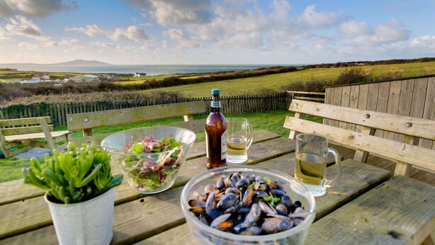 Een houten tafel met een salade, mosselen, een biertje en een glas bier, met op de achtergrond een weids uitzicht over een grasveld en de zee.