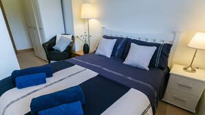 Een net opgemaakt bed met blauw-witte beddengoed, een nachtkastje met een lamp en een stoel met een kussen.