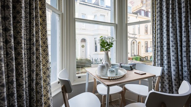 A dining area with a table set for four, a vase with flowers, and a view of the city through large windows. - 2 Sussex Place (Plymouth)